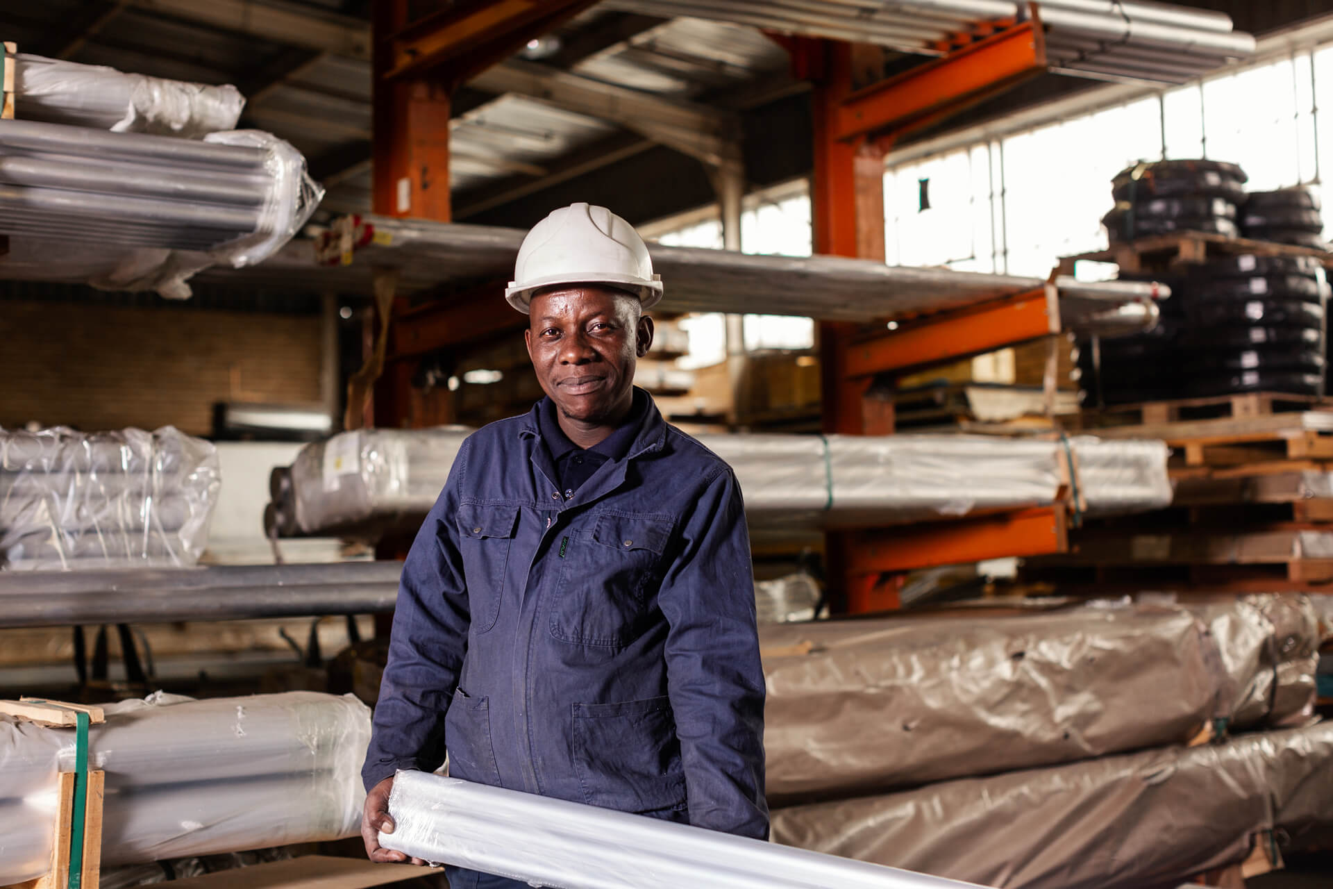 A worker in a hard hat smiles while holding a metal pipe in a warehouse filled with stacked materials.