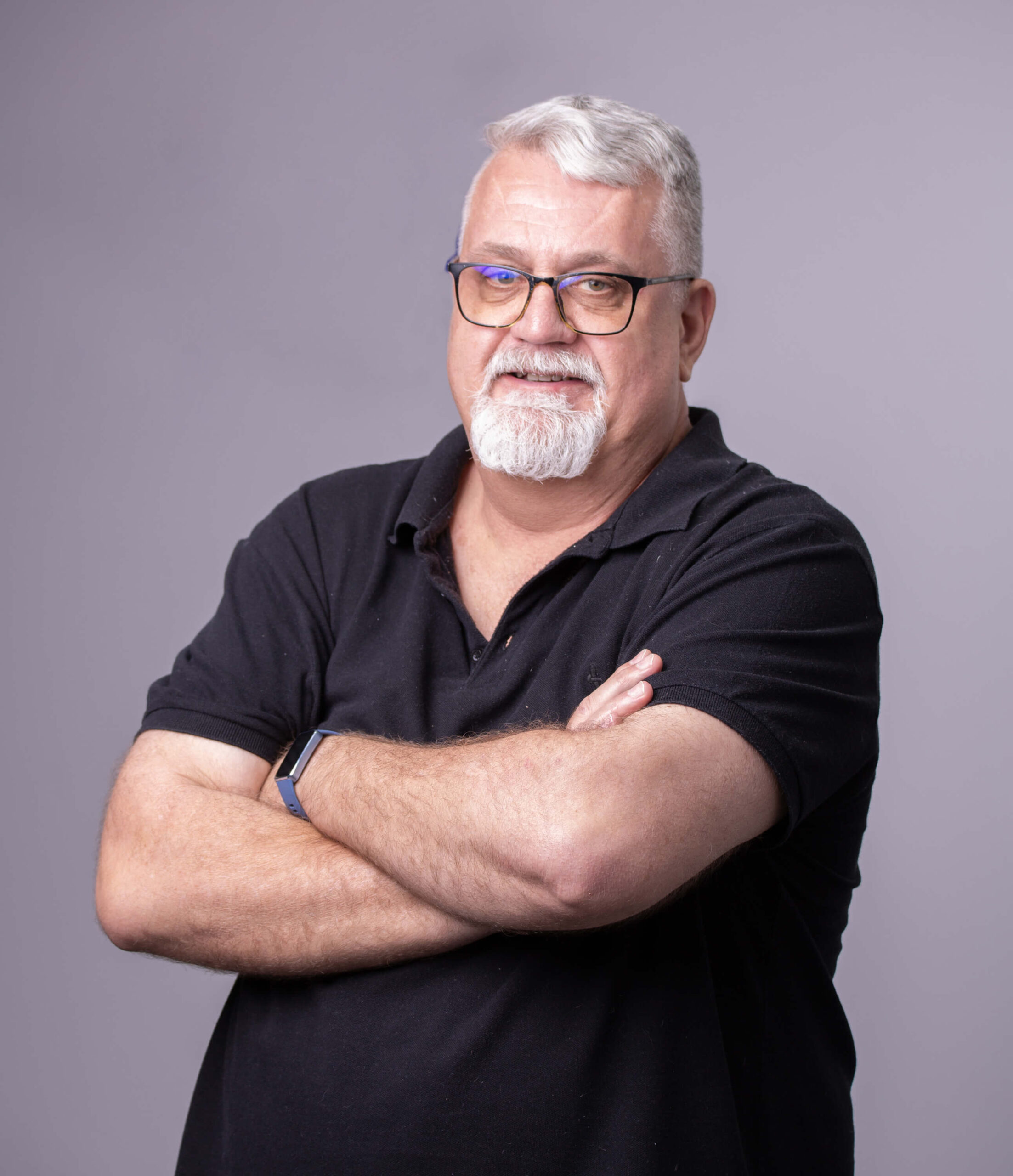 A man with gray hair and glasses smiles while standing with arms crossed against a plain gray background.
