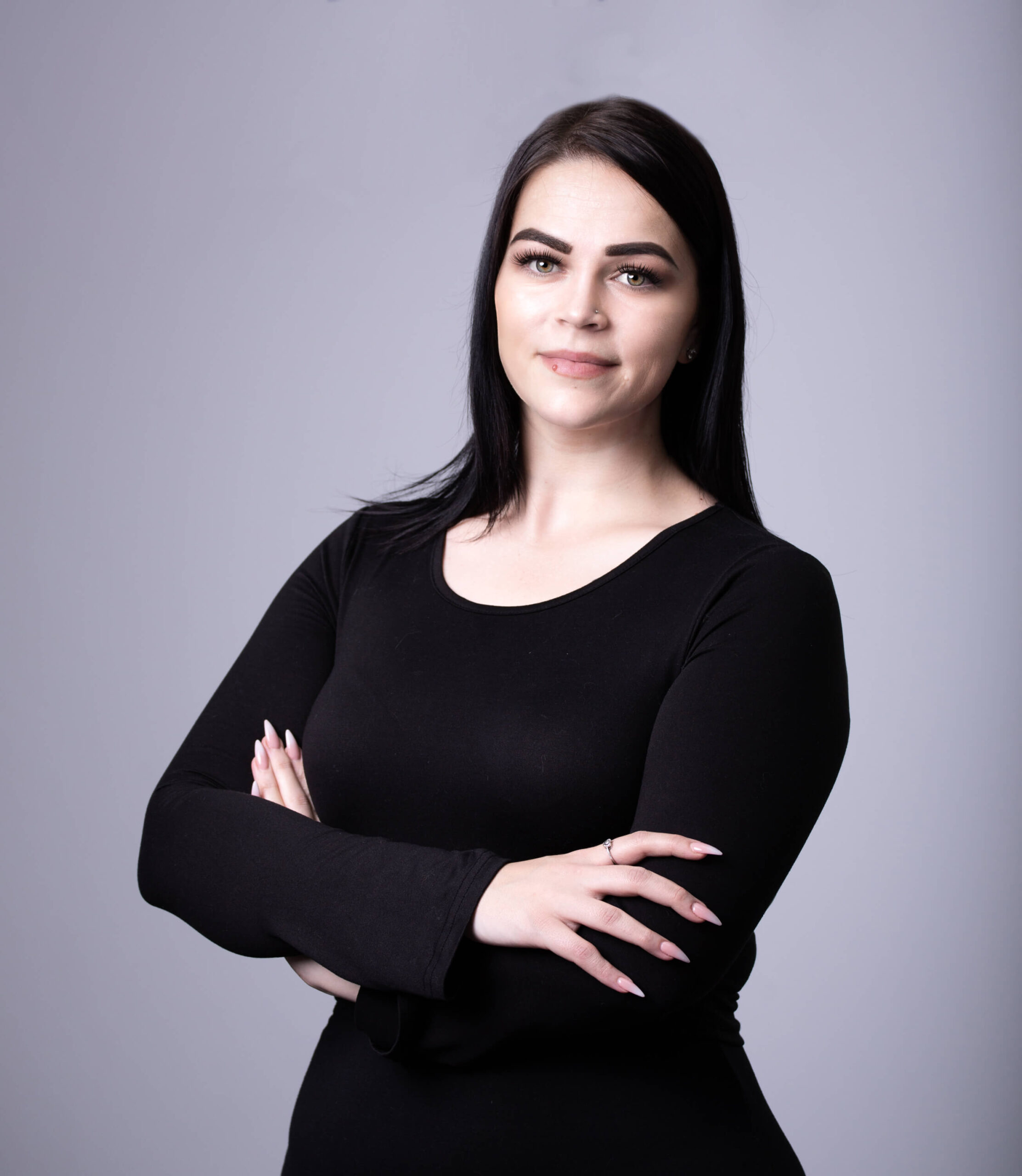 A woman in a black long-sleeve top stands confidently with arms crossed, smiling softly against a gray background.